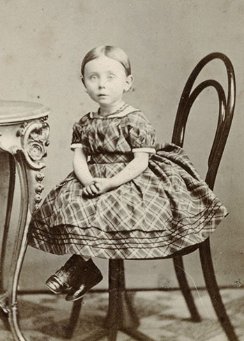 Den lille pige fra den velhavende købmandsfamilie Bay i Randers er klædt i en krinolinekjole som en voksen kvinde. Det er kun kjolelængden, der adskiller pigens kjole fra voksenmoden. Fotografiet er taget i midten af 1860’erne i Randers.