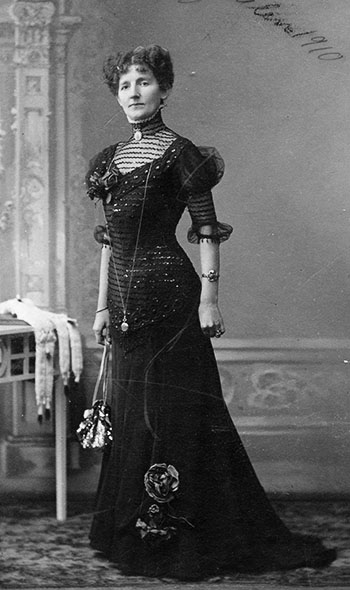 Raffineret dame fotograferet i selskabskjole med slæb i 1910 i Randers. Kjolen, der er S-formet, har besætninger af tylsbroderi i halsudskæringen og tylsærmer. Bemærk stolaen i pelsværk på bordet.